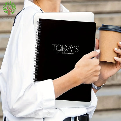Person holding a black planner and a coffee cup, with a blurred background